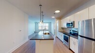 This well-appointed two-bedroom residence offers a thoughtfully designed single-floor layout that balances modern aesthetics with everyday functionality. The open-concept living and kitchen area serves as the heart of the home, anchored by a central island with a dark countertop and pendant lighting overhead. The kitchen is outfitted with crisp white cabinetry, grey subway tile backsplash, dark countertops, and a full suite of stainless steel appliances including a range, microwave, and refrigerator. Light wood-style flooring flows throughout the main living areas, enhancing the bright, airy atmosphere created by large windows that invite generous natural light. The spacious living area is served by a ceiling fan and offers an open sightline to the kitchen. Both bedrooms feature plush carpeting, ceiling fans with integrated lighting, and windows framing urban views. One bedroom includes direct access to a private balcony. The unit is complemented by two bathrooms—a full bath featuring a white vanity with dark countertop, tub/shower combo, and light wood-style flooring, as well as a second bathroom with a single-sink vanity and modern fixtures throughout. Clean white walls and a neutral palette create a cohesive, contemporary feel across the entire residence.