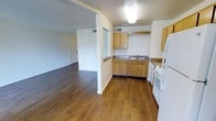 This single-floor space offers a practical and comfortable layout designed for everyday living. The kitchen is equipped with light wood cabinetry providing ample storage, a double-basin sink, dark countertops, and a white range, giving it a clean and functional character. A half-wall partition separates the kitchen from the adjacent living area, creating an open-concept feel while maintaining a sense of definition between spaces. The living area is generously sized, benefiting from the open sightlines that extend toward the entryway, making the space feel bright and connected. Warm hardwood-style flooring runs continuously throughout the kitchen and living areas, adding a cohesive and inviting aesthetic. The ceilings feature a textured finish, and recessed ventilation is visible throughout, supporting a well-ventilated interior. A panel-style entry door is visible from the living area, offering convenient access. Overall, the space presents a straightforward, well-maintained interior with a neutral color palette that provides a versatile backdrop for a variety of furnishing and décor styles.