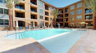 This outdoor amenity space is set within a beautifully designed Mediterranean-inspired complex, featuring warm terra cotta and ochre-toned stucco facades that rise several stories above the central courtyard. The centerpiece of the space is a generously sized swimming pool with crystal-clear turquoise water, surrounded by travertine-style pool decking that provides ample room for lounging. Blue lounge chairs are arranged around the pool's perimeter, offering comfortable spots to relax in the abundant natural sunlight. Tall, mature palm trees are strategically placed throughout the courtyard, adding a resort-like ambiance and providing natural shade. A white pergola structure extends along one side of the pool area, creating a shaded seating zone with additional lounge furniture. Water features, including decorative fountains along the pool's edge, add a dynamic and refreshing element to the setting. A dedicated outdoor kitchen area features multiple stainless steel grills set into a granite-topped counter with a cobblestone paver patio, ideal for outdoor entertaining. The entire space is enclosed by the surrounding multi-story building, creating a private, sheltered atmosphere while maintaining an open, airy feel under clear blue skies.