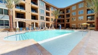 This outdoor amenity space is set within a beautifully designed Mediterranean-inspired complex, featuring warm terra cotta and ochre-toned stucco facades that rise several stories above the central courtyard. The centerpiece of the space is a generously sized swimming pool with crystal-clear turquoise water, surrounded by travertine-style pool decking that provides ample room for lounging. Blue lounge chairs are arranged around the pool's perimeter, offering comfortable spots to relax in the abundant natural sunlight. Tall, mature palm trees are strategically placed throughout the courtyard, adding a resort-like ambiance and providing natural shade. A white pergola structure extends along one side of the pool area, creating a shaded seating zone with additional lounge furniture. Water features, including decorative fountains along the pool's edge, add a dynamic and refreshing element to the setting. A dedicated outdoor kitchen area features multiple stainless steel grills set into a granite-topped counter with a cobblestone paver patio, ideal for outdoor entertaining. The entire space is enclosed by the surrounding multi-story building, creating a private, sheltered atmosphere while maintaining an open, airy feel under clear blue skies.