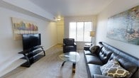 This single-floor space offers a clean, modern aesthetic with a thoughtfully arranged living area as its centerpiece. The living room is outfitted with a tufted black leather sofa and matching armchair, complemented by a sleek oval glass coffee table that adds an elegant touch to the room. A large flat-screen television is mounted on a contemporary media stand along one wall, creating a dedicated entertainment area. Natural light filters into the space through a broad window fitted with horizontal blinds, lending the room a bright and airy feel. Neutral-toned carpeting runs throughout, contributing to the warm and inviting ambiance. The walls are painted in a light, neutral hue, providing a versatile backdrop that enhances the sense of openness. Decorative artwork adds a subtle pop of color to the otherwise understated palette. A hallway visible from the living area leads to additional rooms, with a closet door providing practical storage access. Recessed lighting and a wall-mounted light fixture ensure the space is well-lit throughout the day and evening. Overall, the space presents a comfortable and cohesive environment that balances modern style with everyday functionality.
