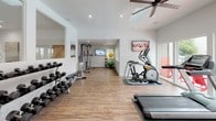 This single-floor fitness center offers a bright, modern environment designed to support a comprehensive workout experience. The space is finished with durable wood-look flooring throughout and features crisp white walls that create a clean, open atmosphere. Large floor-to-ceiling sliding glass doors and expansive windows line one side of the facility, flooding the interior with natural light and offering direct views of an outdoor pool and lounge area complete with red Adirondack chairs and red-umbrella shaded seating.

The cardio zone is positioned near the windows and includes treadmills, an elliptical trainer, and a stationary bike, allowing users to enjoy the outdoor scenery while exercising. Ceiling fans with recessed lighting overhead provide comfortable air circulation throughout the space.

A full-length wall mirror visually expands the room and enhances the sense of openness. The strength training area features a well-stocked dumbbell rack, a flat bench, and a multi-function cable machine, catering to a range of resistance training needs. Medicine balls and stability balls are also available for functional fitness exercises. Mounted flat-screen TVs are positioned for easy viewing from multiple workout zones. A separate room with a glass door, visible at one end, appears to offer additional private fitness space, rounding out this versatile and well-appointed facility.