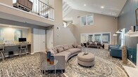Located at 3550 Pleasant Hill Rd in Duluth, Georgia, this well-appointed community lounge space combines modern design with functional versatility. The room is immediately distinguished by its soaring vaulted ceilings, which lend an open, airy quality throughout. A dramatic staircase with an upper-level overlook adds architectural depth, while large windows fitted with blinds invite natural light and views of the surrounding exterior.

The living area serves as the centerpiece, anchored by a curved tufted sectional sofa and complemented by additional seating arranged across patterned area rugs. A striking teal accent wall frames a white marble fireplace with a mounted television above, creating a cozy yet contemporary focal point. An arc floor lamp and coordinating armchair complete this lounge corner with refined detail.

Adjacent to the main lounge, a casual dining area features a high-top bar table with swivel stools, positioned near a window with natural views. A dedicated workstation area offers a built-in granite-topped counter with bar seating and recessed lighting, providing a practical space for business tasks. Aqua-toned artwork adorns the walls of this nook, adding a splash of color. Consistent patterned carpeting flows throughout, unifying the space with a cohesive, sophisticated aesthetic.