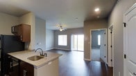 This single-floor residence showcases a thoughtfully designed open-concept layout that seamlessly connects the kitchen and living areas. The kitchen features granite countertops, a stainless steel undermount sink with a dark oil-rubbed bronze faucet, and dark cabinetry, creating a refined and functional workspace. The countertop flows into a breakfast bar that opens directly to the living area, ideal for entertaining or casual dining.

Rich, dark hardwood flooring extends throughout the main living space, providing a warm and elegant foundation that contrasts beautifully with the neutral gray walls. The living area is generously proportioned and is anchored by a ceiling fan, offering both comfort and style. A large sliding glass door floods the space with natural light and provides access to an outdoor area, while an additional window enhances the bright, airy atmosphere.

A doorway visible from the main living area leads to a carpeted room, suggesting a private bedroom or secondary living space. Recessed lighting and smooth ceilings complete the clean, modern aesthetic. Overall, the space presents a cohesive, move-in-ready environment that balances contemporary finishes with practical, comfortable living.