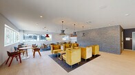 Located at 2600 S Sepulveda Blvd in Los Angeles, this single-floor communal space offers a thoughtfully designed environment that seamlessly blends living and dining functions. The open-plan layout is generously sized, providing a comfortable and versatile gathering area.

The living lounge zone is anchored by a set of plush golden-yellow velvet armchairs arranged around low wooden side tables, all positioned atop a dark area rug. Complementing this seating cluster are sleek charcoal-gray lounge chairs with natural wood legs, paired with deep red accent tables and illuminated by articulating black wall sconces. The walls are painted in a soft sage green, contributing to a calm and inviting atmosphere.

A bold exposed dark brick wall serves as a striking architectural focal point, dividing the space and adding textural contrast to the otherwise clean, white-painted surfaces. Light large-format porcelain tile flooring runs throughout, lending a bright and polished foundation to the room.

The dining area features round and rectangular tables in warm wood and bold burgundy tones, surrounded by contemporary wood-and-fabric chairs. Oversized horizontal windows line the walls, framing views of the surrounding Los Angeles streetscape and flooding the interior with abundant natural light. Statement pendant lights in muted red and gray tones hang overhead, adding a curated, design-forward touch to the overall aesthetic.