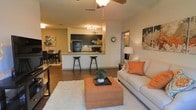 This single-story residence is defined by its open-concept layout, where the living, dining, and kitchen areas flow naturally into one another, creating a cohesive and functional living environment. The living room is anchored by a plush area rug and features a comfortable sofa accented with warm orange throw pillows, complemented by a large upholstered ottoman that doubles as a coffee table. A ceiling fan overhead provides both airflow and ambient lighting to the space. The kitchen is positioned centrally and features dark cabinetry with a tile backsplash, a raised breakfast bar with bar stool seating, and dark appliances that contrast cleanly against light countertops. Track lighting illuminates the kitchen workspace effectively. The dining area is situated just beyond the kitchen, furnished with a dark dining table and chairs, ideal for everyday meals or entertaining. Hardwood-style flooring runs throughout the main areas, adding warmth and visual continuity. The neutral wall tones, warm lighting, and cohesive color palette of oranges and creams give the space an inviting and well-curated ambiance.