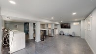 This thoughtfully designed shared laundry facility combines functionality with contemporary aesthetics across a spacious, open floor plan. Large-format gray tile flooring extends throughout, lending a clean, cohesive look to the entire space. Structural support columns are integrated seamlessly into the layout, dividing the room into distinct functional zones without disrupting the open feel.

The laundry area is equipped with a row of full-sized washers and dryers lining one wall, accented by a deep teal ceiling with decorative pendant lighting overhead. A built-in white shelf serves as a dedicated "Charge Up" station, providing convenient device charging while laundry runs. Framed graphic artwork depicting laundry-related icons — detergent, washing machine, and iron — add a playful, polished touch to the walls.

A large folding table with a sleek black metal base occupies the central area, accompanied by a rolling laundry cart for added convenience. Adjacent to the machines, a relaxed waiting area features a wall-mounted flat-screen television, two modern sculptural lounge chairs, and a small ottoman, creating a comfortable space to wait. Recessed lighting throughout ensures bright, even illumination, while small windows allow a modest amount of natural light to filter in.
