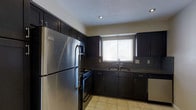 This single-floor space is designed with functionality and style in mind. The kitchen serves as a standout feature, showcasing rich, dark espresso cabinetry that extends from floor to ceiling, offering generous storage throughout. Complementing the cabinetry are dark granite countertops with a subtle speckled pattern, providing both durability and visual appeal. The backsplash coordinates seamlessly with the countertop material, contributing to a cohesive and polished aesthetic. Stainless steel appliances, including a full-size refrigerator, a gas range with overhead microwave, and a dishwasher, bring a modern, professional feel to the cooking space. A double-basin sink is positioned beneath a window fitted with white horizontal blinds, allowing natural light to filter into the room while maintaining privacy. Light-toned tile flooring grounds the darker upper elements, creating a balanced contrast throughout the kitchen. Recessed lighting in the ceiling ensures the space is well-lit, enhancing both the practical cooking environment and the overall ambiance. The layout is efficiently arranged in a U-shape, maximizing counter and cabinet space while maintaining a comfortable workflow for everyday use.