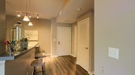 This single-floor residence offers a thoughtfully designed layout that balances functionality and comfort. The entryway makes a strong first impression with rich, dark hardwood-style flooring that extends throughout the space, complemented by neutral greige wall tones that create a warm and inviting atmosphere.

The entry corridor leads to the main living areas, where soft ambient lighting from track-mounted and pendant fixtures enhances the overall warmth of the interior. White paneled doors provide clean, classic detailing throughout, including a louvered closet door that adds a touch of traditional character while offering practical storage access.

The open transition between the entryway and the kitchen or dining area is highlighted by a curved counter edge and bar-style seating with contemporary metal stools, suggesting a casual and social dining setup. Overhead, the smooth ceiling features recessed HVAC vents and a smoke detector, reflecting a well-maintained and code-compliant interior.

The muted color palette, consistent flooring, and careful lighting choices throughout give the space a cohesive and polished feel. The overall design leans toward modern simplicity while incorporating warm textures and classic architectural details, making it a versatile and livable environment.