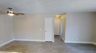 This single-floor residential space presents a clean, functional layout with a consistent design aesthetic throughout. The interior is finished with light wood-look laminate or vinyl plank flooring that runs seamlessly across the main living area and into the connecting hallway, providing a cohesive and easy-to-maintain surface underfoot. The walls are painted in a warm, neutral beige tone that complements the light flooring and contributes to a bright, open feel throughout the space. Overhead, a textured popcorn ceiling adds a classic residential character, with flush-mount ceiling fixtures providing straightforward overhead lighting. The main living area features a white interior door providing access to what appears to be a closet, adding convenient storage within the space. A hallway leads from the main area toward additional rooms, with doorways visible at the end suggesting access to a bedroom and potentially a bathroom, both finished in the same neutral palette. Crisp white baseboards frame each room, adding a polished contrast to the beige walls and light flooring. The overall ambiance is understated and neutral, making the space highly adaptable to a variety of interior styles and furnishing preferences.