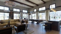 This stylish single-floor space makes a strong architectural impression with its dramatic high ceilings accented by rich, dark exposed wood beams that run the length of the room. Abundant natural light pours in through large floor-to-ceiling windows and elevated clerestory windows positioned along the upper walls, creating a bright and airy atmosphere while offering views of the surrounding neighborhood. The main living area is anchored by a plush sectional sofa and leather accent chairs arranged around a round coffee table, all set atop a soft area rug over wide-plank hardwood flooring. A sleek pool table takes center stage in the open layout, making the space well-suited for social gatherings and recreation. Bar-height seating and a countertop station along one side provide additional gathering space. Contemporary pendant lighting and a sculptural chandelier complement the recessed ceiling lights, adding warmth and visual interest to the expansive interior. Neutral gray walls and crisp white drapery panels tie the design together, lending a clean, modern aesthetic throughout. The open floor plan allows for seamless flow between the lounge, game, and casual seating areas, making this a versatile and inviting communal space.