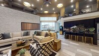 This expansive amenity lounge is designed to serve as a dynamic social hub, combining multiple functional areas within a single, open-concept floor plan. Upon entry through wide glass doors, guests are welcomed by large-format tile flooring and soaring double-height ceilings that lend the space a grand, airy quality. A mezzanine-level overlook with metal railings adds architectural depth and visual interest overhead.

The central lounge area is anchored by a dramatic floor-to-ceiling stacked stone accent wall, against which a large flat-screen display is mounted. Plush neutral sofas and eclectic patterned accent chairs are arranged to encourage conversation. Recessed lighting and pendant fixtures provide layered illumination throughout.

Adjacent to the lounge, a well-appointed kitchenette features dark cabinetry, granite countertops, a stone tile backsplash, and a dedicated coffee station. A bar-height counter with seating provides an informal gathering spot nearby.

A dining area with a dark wood table sits beside floor-to-ceiling windows dressed in patterned drapes, offering views of an outdoor patio with covered seating. The 3D floorplan also reveals a billiards table, rounding out the entertainment offerings. The overall ambiance is contemporary and refined.