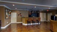 This single-floor space is designed with a warm, inviting aesthetic anchored by a central bar or kitchenette area that serves as the room's focal point. The bar counter features rich, decorative detailing along its base, complemented by dark cabinetry and a granite or stone-finished countertop. Four upholstered bar stools in warm brown tones line the counter, providing comfortable seating for socializing or entertaining.

Overhead, a trio of pendant lights with dark metal finishes hang at varying heights, adding visual interest and focused illumination to the bar area. Recessed lighting throughout the ceiling ensures the space remains well-lit overall. Crown molding along the ceiling perimeter adds a touch of architectural refinement.

The flooring consists of large-format tiles in warm amber and terracotta hues, contributing to the earthy color palette that defines the space. The walls are finished in a complementary taupe tone, creating a cohesive and grounded atmosphere. Two standard doors on either side of the space provide access to adjoining areas. A small piece of abstract wall art adds a decorative accent, rounding out the room's well-curated, functional design.