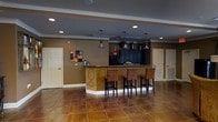 This single-floor space is designed with a warm, inviting aesthetic anchored by a central bar or kitchenette area that serves as the room's focal point. The bar counter features rich, decorative detailing along its base, complemented by dark cabinetry and a granite or stone-finished countertop. Four upholstered bar stools in warm brown tones line the counter, providing comfortable seating for socializing or entertaining.

Overhead, a trio of pendant lights with dark metal finishes hang at varying heights, adding visual interest and focused illumination to the bar area. Recessed lighting throughout the ceiling ensures the space remains well-lit overall. Crown molding along the ceiling perimeter adds a touch of architectural refinement.

The flooring consists of large-format tiles in warm amber and terracotta hues, contributing to the earthy color palette that defines the space. The walls are finished in a complementary taupe tone, creating a cohesive and grounded atmosphere. Two standard doors on either side of the space provide access to adjoining areas. A small piece of abstract wall art adds a decorative accent, rounding out the room's well-curated, functional design.
