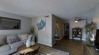 This single-story space showcases a thoughtfully arranged interior with a cohesive, neutral color palette throughout. The main living area features dark hardwood-style flooring that contrasts cleanly against the light gray walls, creating a warm yet contemporary aesthetic. A textured popcorn ceiling adds dimension overhead, while HVAC vents are discreetly integrated into the ceiling for climate control.

The living space is anchored by a comfortable seating arrangement and a round wooden coffee table, complemented by decorative accents including ceramic vases and a vibrant botanical wall art piece featuring blue and gold tones. A tall decorative vase rests atop a small accent table near a partial wall that defines the space.

Beyond the main living area, a hallway leads to additional rooms, with a glimpse of what appears to be a kitchen with warm-toned cabinetry visible in the background. A ceiling fan with an integrated light fixture is positioned in an adjacent area, ensuring comfortable airflow. A wooden bookcase or display cabinet along the far wall adds functional storage. Natural light filters into the space, enhancing the inviting and well-maintained ambiance throughout.