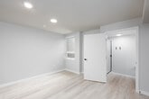 This single-floor unit presents a thoughtfully designed layout that maximizes functionality within a compact footprint. The interior is finished with light-toned hardwood-style flooring throughout, contributing to a bright, cohesive aesthetic across all areas of the space.

The kitchenette is a standout feature, equipped with natural wood-grain cabinetry accented by matte black hardware, a grey stone-look countertop, an undermount stainless steel sink with a black faucet, a built-in electric cooktop, an over-the-counter microwave, and a compact stainless steel refrigerator. Upper and lower cabinets provide practical storage, making efficient use of the available wall space.

The main living area is open and versatile, with neutral warm-beige walls, clean white baseboards, and a smoke detector installed on the smooth white ceiling. A window along one wall allows natural light to filter into the room, creating an airy atmosphere.

The bathroom is finished in the same neutral tones as the rest of the unit, featuring a modern toilet and an oval white-framed mirror mounted above it, adding a decorative touch to the otherwise minimalist space. The overall design of the unit is modern and move-in ready, with consistent finishes that lend a clean, unified appearance throughout.