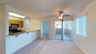This single-floor residential unit offers a practical and comfortable layout designed to maximize open living space. The main living area is carpeted and finished with a textured ceiling, lending a warm and cozy feel throughout. A ceiling fan with integrated lighting serves as both a functional and central design element in the living room, ensuring year-round comfort. Large sliding glass doors open directly onto a private balcony, allowing natural light to flow into the living space while offering outdoor views of the surrounding greenery.

The kitchen is positioned adjacent to the living area, separated by a raised breakfast bar counter that allows for an open-concept connection between the two spaces. Warm-toned cabinetry and pendant lighting above the counter give the kitchen a classic, inviting character, while a window provides additional natural light. A doorway from the living area leads into a secondary room, also carpeted, providing flexible use as a bedroom or additional living space.

The overall design of the unit is straightforward and functional, with neutral tones throughout that create a clean, versatile backdrop. The combination of indoor comfort and outdoor balcony access makes this a well-rounded residential space suited for everyday living.