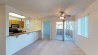 This single-floor residential unit offers a practical and comfortable layout designed to maximize open living space. The main living area is carpeted and finished with a textured ceiling, lending a warm and cozy feel throughout. A ceiling fan with integrated lighting serves as both a functional and central design element in the living room, ensuring year-round comfort. Large sliding glass doors open directly onto a private balcony, allowing natural light to flow into the living space while offering outdoor views of the surrounding greenery.

The kitchen is positioned adjacent to the living area, separated by a raised breakfast bar counter that allows for an open-concept connection between the two spaces. Warm-toned cabinetry and pendant lighting above the counter give the kitchen a classic, inviting character, while a window provides additional natural light. A doorway from the living area leads into a secondary room, also carpeted, providing flexible use as a bedroom or additional living space.

The overall design of the unit is straightforward and functional, with neutral tones throughout that create a clean, versatile backdrop. The combination of indoor comfort and outdoor balcony access makes this a well-rounded residential space suited for everyday living.