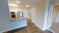 This single-floor residence showcases a thoughtfully designed open-concept layout that seamlessly connects the kitchen and living areas. The kitchen serves as a functional centerpiece, featuring a large island with striking granite countertops and an integrated sink, providing ample workspace and casual dining potential. Stainless steel appliances, including a refrigerator, microwave, and range, are complemented by crisp white cabinetry that extends to the upper walls, offering generous storage throughout.

The flooring throughout the space consists of rich, dark hardwood-style planks that add warmth and visual continuity from room to room. Neutral, light beige walls create a bright and welcoming atmosphere, enhanced by recessed and flush-mount ceiling lighting that ensures consistent illumination across the space.

A hallway visible from the kitchen leads to additional rooms, flanked by clean white six-panel doors that contribute to the home's cohesive, modern aesthetic. The open sightlines and uncluttered design give the space an airy, organized feel. Overall, the unit presents a clean, contemporary style well-suited for comfortable everyday living.