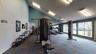 This single-floor fitness center offers a comprehensive workout environment designed to accommodate a variety of exercise routines. The space is divided into distinct functional zones, each dedicated to a specific type of training. The walls are painted in a two-tone scheme of teal and soft light blue, creating an energetic yet focused atmosphere, while motivational phrases such as "Just Do It," "Beast Mode," and "It's Simple… DO IT!" are displayed throughout to encourage users.

The cardio zone features multiple treadmills and stationary bikes positioned near large windows that provide views of the surrounding outdoor greenery, allowing natural light to brighten the space. Overhead fluorescent strip lighting ensures consistent illumination throughout. A full-length mirror spans one wall, visually expanding the room and aiding form checks during workouts.

The strength training area includes a multi-station cable machine with an adjustable bench, a wall-mounted weight rack stocked with a range of dumbbells, and additional cable resistance equipment. Framed fitness posters and informational signage, including a safety checklist, are mounted on the walls.

The flooring throughout consists of patterned carpet tiles in muted blue and gray tones, providing cushioning underfoot. French doors connect to an adjacent room, and exterior-facing doors with large glass panels offer easy access and reinforce the bright, open feel of the facility.