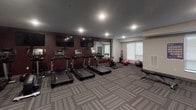 Located at 2180 Highway 13 in Mendota Heights, Minnesota, this fitness center offers a well-appointed, single-floor workout facility designed to support a comprehensive exercise routine. The space features a bold interior palette of deep burgundy and charcoal gray, with patterned carpet tiles underfoot and recessed lighting throughout that creates a focused, energetic atmosphere.

The cardio zone is anchored by a row of Life Fitness treadmills, each equipped with an individual display screen. Multiple flat-screen TVs are mounted along the burgundy accent wall, allowing users to stay entertained during workouts. A spin bike rounds out the cardio offerings. Full-length mirrors line one wall, visually expanding the space and providing form-checking functionality.

The strength training area includes a tiered dumbbell rack with a broad range of free weights, a flat weight bench, and a multi-station cable machine for a variety of resistance exercises. Stability and medicine balls are neatly stored along the wall, adding versatility to workout options.

Natural light filters in through large windows on one side of the room, complementing the recessed overhead lighting. A water fountain and hydration station are conveniently positioned within the space, along with access doors that connect to additional building amenities. The overall layout is open and functional, providing ample room for movement and exercise.