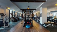 This fitness center is thoughtfully designed across a single level, offering a comprehensive range of workout zones within a stylish, boutique-inspired environment. The space is defined by rich hardwood-style flooring, exposed dark wood ceiling beams, and textured white brick accent walls that lend warmth and character throughout.

The cardio zone runs along a wall of floor-to-ceiling windows, flooding the area with natural light and providing open views of an outdoor terrace. Ellipticals, stair climbers, treadmills, and a rowing machine are arranged to take full advantage of these views. Overhead, large ceiling fans and warm pendant lights maintain a comfortable ambiance.

A dedicated cycling studio features smart Echelon bikes equipped with individual screens, arched mirrors, and a Reflect interactive display for on-demand classes. Adjacent areas are equipped with cable machines, multi-functional strength stations, medicine ball racks, and a BOSU trainer, catering to functional and resistance training needs.

A mirrored stretching and movement area with a barre rail offers space for flexibility work or group instruction. Built-in storage cabinetry and curated motivational signage enhance the polished, intentional aesthetic. A water station and sauna access further support recovery and wellness.