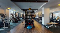 This fitness center is thoughtfully designed across a single level, offering a comprehensive range of workout zones within a stylish, boutique-inspired environment. The space is defined by rich hardwood-style flooring, exposed dark wood ceiling beams, and textured white brick accent walls that lend warmth and character throughout.

The cardio zone runs along a wall of floor-to-ceiling windows, flooding the area with natural light and providing open views of an outdoor terrace. Ellipticals, stair climbers, treadmills, and a rowing machine are arranged to take full advantage of these views. Overhead, large ceiling fans and warm pendant lights maintain a comfortable ambiance.

A dedicated cycling studio features smart Echelon bikes equipped with individual screens, arched mirrors, and a Reflect interactive display for on-demand classes. Adjacent areas are equipped with cable machines, multi-functional strength stations, medicine ball racks, and a BOSU trainer, catering to functional and resistance training needs.

A mirrored stretching and movement area with a barre rail offers space for flexibility work or group instruction. Built-in storage cabinetry and curated motivational signage enhance the polished, intentional aesthetic. A water station and sauna access further support recovery and wellness.