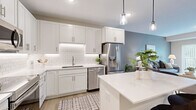 This well-appointed one-bedroom, one-bathroom residence in Lakeville, MN offers a smart, efficient layout across a single floor. The open-concept kitchen and living area serves as the heart of the home, where crisp white shaker cabinetry, white subway tile backsplash, and quartz countertops give the kitchen a clean, contemporary look. Stainless steel appliances — including a refrigerator with ice maker, range, microwave, and dishwasher — provide full functionality, while pendant lighting and under-cabinet lighting add a polished finish. A kitchen island with seating creates a natural gathering point that flows into the living room. The living area is bright and welcoming, featuring light wood-style flooring and a full sliding glass door with vertical blinds that opens to an outdoor view, filling the space with natural light. The bedroom offers plush carpeting, a large window overlooking greenery, and access to a generous walk-in closet fitted with wire shelving. The bathroom features a white vanity with a quartz countertop and wood-style flooring. Neutral tones and modern finishes throughout lend the entire residence a cohesive, contemporary ambiance.