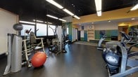 Located at 808 Memorial Dr in Cambridge, MA, this single-floor fitness center offers a well-equipped workout environment designed to support a range of exercise routines. The main workout area features durable rubber flooring and is lined with a variety of cardio machines, including treadmills, ellipticals, and stationary bikes, as well as multi-use strength training cable machines and free accessories such as stability balls and exercise mats. Overhead, long fluorescent strip lighting suspended from a dark-painted ceiling provides ample illumination throughout the space. Wall-mounted mirrors and informational fitness posters contribute to both functionality and visual openness. A dedicated locker and changing area transitions the space from the workout floor, featuring colorful wood-paneled lockers in shades of blue and green, a built-in shelving unit, and a cushioned bench — adding a welcoming, organized feel. Windows along the perimeter allow natural light to filter in, offering views of the surrounding outdoor landscape. A wall-mounted television is positioned for visibility during workouts, and the open layout ensures comfortable circulation between equipment zones, making this fitness center a practical and functional facility.