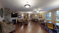 This single-story communal facility is thoughtfully designed to provide a warm and functional environment across several distinct areas. The centerpiece of the space is a generously sized common room featuring rich hardwood flooring, multiple ceiling fans with light fixtures, and pendant lighting that illuminates the open layout. A striking stone-accented fireplace anchors one end of the room, paired with a wall-mounted flat-screen television and comfortable upholstered seating arrangements. Framed artwork and wall sconces add a refined yet approachable aesthetic throughout.

Adjacent to the lounge area is a dedicated dining space outfitted with dark wood tables and cushioned chairs. Large windows overlook an outdoor area, filling the room with natural light and exterior views. A second dining area mirrors this setup, offering additional seating capacity.

The facility includes a well-equipped kitchen featuring granite countertops, a central island, stainless steel appliances, and light maple cabinetry—functional and neatly appointed for communal use.

The carpeted corridors are wide and accessible, featuring coffered ceiling details, soft wall sconces, decorative framing, and elevator access for convenient navigation. Small seating alcoves are tucked along the hallways, providing intimate spots for quiet conversation or rest.