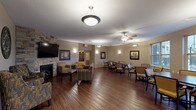 This single-story communal facility is thoughtfully designed to provide a warm and functional environment across several distinct areas. The centerpiece of the space is a generously sized common room featuring rich hardwood flooring, multiple ceiling fans with light fixtures, and pendant lighting that illuminates the open layout. A striking stone-accented fireplace anchors one end of the room, paired with a wall-mounted flat-screen television and comfortable upholstered seating arrangements. Framed artwork and wall sconces add a refined yet approachable aesthetic throughout.

Adjacent to the lounge area is a dedicated dining space outfitted with dark wood tables and cushioned chairs. Large windows overlook an outdoor area, filling the room with natural light and exterior views. A second dining area mirrors this setup, offering additional seating capacity.

The facility includes a well-equipped kitchen featuring granite countertops, a central island, stainless steel appliances, and light maple cabinetry—functional and neatly appointed for communal use.

The carpeted corridors are wide and accessible, featuring coffered ceiling details, soft wall sconces, decorative framing, and elevator access for convenient navigation. Small seating alcoves are tucked along the hallways, providing intimate spots for quiet conversation or rest.