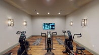 Located on N Pecan Pkwy in Justin, TX, this single-floor fitness facility is thoughtfully designed to provide a focused and polished workout environment. The space is divided into distinct zones connected by black-framed glass doors and partitions, lending the interior a modern, studio-like aesthetic while maintaining an open, airy feel throughout.

The main exercise room serves as the heart of the facility, housing several stationary and air resistance bikes arranged on patterned area rugs atop warm hardwood-style flooring. A large wall-mounted flat-screen television anchors the front wall, allowing users to follow virtual fitness classes or training content during their sessions. Elegant cross-pattern wall sconces on either side provide warm ambient lighting, complemented by recessed ceiling fixtures that evenly illuminate the room.

An adjoining area functions as a transitional space and entry zone, featuring a wooden bench for convenience and rack-style storage. Black-framed floor-to-ceiling glass walls provide clear sightlines between rooms. A dedicated virtual fitness studio room, labeled accordingly on its glass door, offers additional workout space with more bikes and patterned floor mats.

The entrance area features a striking sputnik chandelier and glass exterior doors, welcoming visitors with a refined, upscale touch that carries throughout the entire facility.