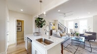 This two-story home in Gentry, Arkansas, is designed with a clean, contemporary aesthetic characterized by neutral tones, light hardwood flooring, and an abundance of natural light throughout. The main floor centers on an open-concept layout where the kitchen flows seamlessly into the living area. The kitchen is outfitted with white shaker cabinetry, quartz countertops, stainless steel appliances including a French door refrigerator and electric range, a subway tile backsplash, and a central island with an undermount sink and pendant lighting overhead. The adjacent living room features recessed lighting, a ceiling fan, and large windows that invite natural light, with a staircase fitted with modern metal cable railings serving as a visual focal point. A secondary sitting area on the main level provides additional flexible living space. The upper floor hosts multiple bedrooms, each thoughtfully appointed with recessed lighting, ceiling fans, and well-sized windows offering views of the surrounding neighborhood. A shared bathroom on the upper level features a dual-sink vanity with white cabinetry, quartz countertops, and tile flooring. Throughout both floors, the consistent use of soft neutral walls, quality finishes, and modern fixtures creates a cohesive and polished atmosphere.