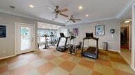 Located in Chesapeake, Virginia, this fitness facility offers a thoughtfully designed space across two interconnected exercise rooms. The layout flows seamlessly between areas, creating an open and spacious feel ideal for a full workout experience.

Both rooms feature soft blue-gray walls, crown molding, and a distinctive checkered carpet in warm terracotta and tan tones, lending the space a clean yet inviting aesthetic. Recessed lighting and multiple ceiling fans with integrated light fixtures ensure the rooms remain well-lit and comfortably ventilated throughout.

The facility is equipped with a comprehensive selection of cardio equipment, including treadmills, elliptical trainers, and stationary bikes from quality brands. Wall-mounted flat-screen televisions are positioned for easy viewing during workouts. Large mirrors in one of the rooms enhance the sense of depth and space while serving a functional purpose for exercise form.

Natural light flows generously through multiple windows and glass-panel doors, offering views of the surrounding outdoor area and glimpsing a nearby pool. A dedicated entry area with tile flooring provides a clean transition into the carpeted exercise zones. Safety features such as fire extinguishers are visibly in place. Decorative artwork on the walls adds a polished, welcoming touch to this practical and well-maintained fitness space.