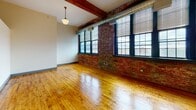 Located at 102 Sprague St in Providence, Rhode Island, this single-floor loft-style unit showcases a compelling blend of industrial heritage and modern comfort. The open-plan living and kitchen area is the centerpiece of the space, characterized by soaring ceilings accented by exposed wooden beams and large, multi-pane black-framed windows that line the brick walls, flooding the interior with generous natural light and offering views of the surrounding neighborhood.

Original hardwood floors in warm honey tones stretch throughout the main area, adding richness and character to the space. The exposed brick walls lend an authentic industrial aesthetic, complementing the airy, loft-like atmosphere. Visible ductwork and pendant lighting fixtures reinforce the converted-building charm.

The kitchen is well-appointed, featuring abundant maple cabinetry, dark countertops with a teal backsplash, and a full suite of white appliances including a gas range, dishwasher, and refrigerator. A pendant light adds a stylistic accent above the workspace.

The bathroom is cleanly finished with a tub-shower combination framed by white tile, a wood-toned vanity with a dark countertop, mosaic tile flooring, and a wall-mounted mirror with overhead lighting, providing a functional and tidy retreat.