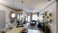 This well-appointed single-floor residence showcases a thoughtfully designed open-concept layout that seamlessly connects the kitchen and living areas. The kitchen features a granite or stone-speckled countertop bar that serves as both a functional workspace and a casual dining surface, complemented by elegant pendant lighting that adds a warm, ambient glow. Crown molding runs along the ceiling, lending a refined architectural touch throughout the space. The living area is anchored by a plush area rug and a contemporary sectional sofa adorned with accent pillows, while a ceiling fan overhead ensures year-round comfort. A large glass door and windows at the far end of the living room flood the space with natural light and provide a view to the outdoors. Hardwood-style flooring runs throughout, adding warmth and continuity to the open floor plan. A bedroom is partially visible through a mirror or open doorway, hinting at additional private living space. Decorative elements, including wall art, lush indoor plants, and stylish light fixtures, contribute to the modern yet comfortable ambiance that defines this residence.