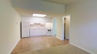 This single-floor space features an open-concept layout that connects the kitchen and living area, creating a seamless flow throughout the interior. The kitchen is outfitted with white cabinetry extending to near ceiling height, white appliances including a refrigerator and electric range, and a single-basin sink positioned beneath upper cabinets. A skylight panel overhead floods the kitchen with natural overhead light, enhancing the bright, airy atmosphere. The countertops and cabinetry maintain a clean, uniform white finish that contributes to the space's light and neutral aesthetic. Light hardwood flooring extends through the kitchen and transitions into a carpeted living area, offering a subtle distinction between the two functional zones. The walls are finished in a soft, warm neutral tone throughout. A doorway off the main area leads to what appears to be a walk-in closet, providing practical storage. The overall ambiance is clean, minimal, and well-lit, with a straightforward layout that maximizes usability across the open floor plan.
