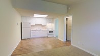 This single-floor space features an open-concept layout that connects the kitchen and living area, creating a seamless flow throughout the interior. The kitchen is outfitted with white cabinetry extending to near ceiling height, white appliances including a refrigerator and electric range, and a single-basin sink positioned beneath upper cabinets. A skylight panel overhead floods the kitchen with natural overhead light, enhancing the bright, airy atmosphere. The countertops and cabinetry maintain a clean, uniform white finish that contributes to the space's light and neutral aesthetic. Light hardwood flooring extends through the kitchen and transitions into a carpeted living area, offering a subtle distinction between the two functional zones. The walls are finished in a soft, warm neutral tone throughout. A doorway off the main area leads to what appears to be a walk-in closet, providing practical storage. The overall ambiance is clean, minimal, and well-lit, with a straightforward layout that maximizes usability across the open floor plan.