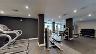 This fitness center is a thoughtfully designed, single-floor facility that combines modern aesthetics with a comprehensive selection of exercise equipment. The space features a striking interior defined by dark charcoal accent walls, bold structural columns, and crisp white ceilings fitted with recessed lighting, creating a dynamic and motivating atmosphere.

The cardio zone is outfitted with a row of Star Trac treadmills and multiple elliptical trainers, positioned to take advantage of large floor-to-ceiling windows that invite natural light and outdoor views, enhancing the workout experience. Rubber flooring in this area provides comfort and durability underfoot.

The strength training section offers a well-rounded selection of equipment, including a multi-station cable and pulley machine, a dedicated bench press and leg roller station, and a full rack of dumbbells neatly organized on tiered shelving. A wall-mounted flat-screen TV serves as both an entertainment and instructional focal point in this zone. Large-format dark tile flooring grounds this area with a sleek, professional finish.

Throughout the facility, strategic use of mirrors amplifies the sense of space and allows users to monitor their form. The overall layout is open and spacious, ensuring ease of movement between zones. The combination of contemporary finishes, abundant equipment variety, and natural light makes this fitness center a well-appointed and functional space.