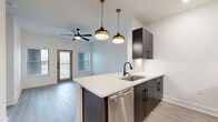 This thoughtfully designed single-floor unit offers a contemporary open-concept layout that maximizes its compact footprint. The kitchen serves as a central focal point, featuring a peninsula with a crisp white quartz countertop, an undermount sink with a matte black faucet, and dark wood-grain cabinetry with matching black hardware. A full suite of stainless steel appliances—including a refrigerator, range, microwave, and dishwasher—adds a polished, modern touch. Two brass-and-black pendant lights hang above the peninsula, complementing the recessed lighting throughout. A wine rack detail adds a refined design element to the upper cabinetry.

The open living area flows naturally from the kitchen, bathed in natural light from a glass-paned entry door and adjacent window. A ceiling fan with integrated lighting serves both areas. Light-toned hardwood-style flooring runs consistently throughout the unit, creating visual continuity and a clean, airy atmosphere.

The bedroom is a well-proportioned private retreat with its own ceiling fan, a large window allowing ample natural light and outdoor views, and the same consistent flooring. A dedicated closet provides practical storage. Crisp white walls throughout the unit enhance the bright, modern ambiance.