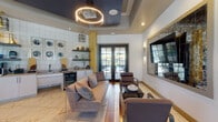 This thoughtfully designed amenity lounge presents a sophisticated blend of contemporary finishes and inviting communal spaces across a single floor. The entrance foyer sets the tone immediately, featuring a bold yellow tray ceiling, large black-framed glass doors, warm wood-plank flooring, and crisp white tile—creating a bright, welcoming transition into the main lounge.

The lounge area is anchored by a plush gray sectional sofa and modern leather armchairs arranged around a sleek coffee table. A large flat-screen television is mounted within a dramatic antique-mirror surround, flanked by elegant wall sconces that add ambient warmth. A statement circular ring chandelier overhead further elevates the space. French doors at the far end connect directly to an outdoor pool area, flooding the lounge with natural light and providing easy indoor-outdoor access.

Adjacent to the lounge, a full wet bar runs along a white subway tile wall, outfitted with white cabinetry, an undermount sink, a built-in beverage cooler, a coffee station, and open shelving displaying decorative accents and glassware. A separate dining nook features dark tables and wood-and-metal chairs positioned alongside large windows with adjustable blinds, offering a quieter setting for work or casual dining. Marble-look tile flooring flows throughout, unifying the space with a polished, resort-inspired aesthetic.