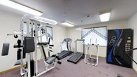 This single-floor fitness room offers a practical and functional space designed to support a variety of workout routines. The room is outfitted with a selection of exercise equipment, including two treadmills for cardiovascular training, a multi-station cable weight machine for strength and resistance exercises, and a standing elliptical-style stepper for low-impact full-body workouts. The flooring is carpeted in a neutral tone, providing a comfortable surface underfoot. Overhead, flush-mounted fluorescent lighting fixtures ensure the room is well-lit throughout. The walls are painted in a light, neutral color, contributing to a clean and open feel. Windows along one wall are dressed with decorative patterned curtains, allowing for natural light while maintaining privacy. A wall-mounted mirror on one side of the room enhances the sense of space and allows users to monitor their form during exercise. The ceiling features a central ventilation unit to support air circulation. Overall, the space is straightforward and purpose-driven, offering residents or guests a convenient and accessible venue for maintaining their fitness routines.