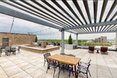 This rooftop terrace is a thoughtfully designed outdoor space that offers an exceptional combination of functional amenities and striking panoramic views. The terrace is finished with large-format stone pavers throughout, providing a clean, cohesive surface across the entire level. A substantial steel pergola serves as the architectural centerpiece, spanning a generous portion of the terrace and outfitted with bistro string lights that contribute to an inviting atmosphere during evening use.

Beneath the pergola, a wooden dining table with ornate cast-iron chairs provides a defined alfresco dining area suited for group gatherings. Adjacent to this is a raised stone fireplace surround, clad in stacked natural stone, creating a focal point for a lounge area complete with sun loungers positioned nearby. Potted evergreen topiaries are strategically placed throughout the terrace, adding vertical greenery and a sense of structure to the open-air layout.

Sleek white metal railings line the perimeter, ensuring unobstructed sightlines to the surrounding environment. Views extend across a nearby athletic field and urban streetscape in one direction, while another vantage point reveals a distant body of water framed by a lush tree canopy. The overall ambiance is that of an elevated, resort-style outdoor retreat.