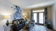 This single-floor residence features two bedrooms and two bathrooms, thoughtfully designed to maximize both comfort and functionality. The living area serves as the centerpiece of the home, showcasing neutral-toned walls that provide a versatile backdrop for décor. A large abstract artwork anchors one wall, adding a bold visual focal point to the space. The room accommodates generous seating arrangements and benefits from a sliding glass door that invites abundant natural light while offering direct access to an outdoor area. Decorative drapery in complementary tones frames the door, enhancing the overall aesthetic. The flooring features a combination of hard surfaces and area rugs, adding warmth and definition to the living space. The layout flows naturally, making efficient use of the available square footage across the single level. With clean lines, ample lighting options including accent lamps, and carefully considered design details, the space strikes a balance between modern style and everyday livability, making it a practical yet visually appealing residence.
