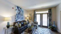 This single-floor residence features two bedrooms and two bathrooms, thoughtfully designed to maximize both comfort and functionality. The living area serves as the centerpiece of the home, showcasing neutral-toned walls that provide a versatile backdrop for décor. A large abstract artwork anchors one wall, adding a bold visual focal point to the space. The room accommodates generous seating arrangements and benefits from a sliding glass door that invites abundant natural light while offering direct access to an outdoor area. Decorative drapery in complementary tones frames the door, enhancing the overall aesthetic. The flooring features a combination of hard surfaces and area rugs, adding warmth and definition to the living space. The layout flows naturally, making efficient use of the available square footage across the single level. With clean lines, ample lighting options including accent lamps, and carefully considered design details, the space strikes a balance between modern style and everyday livability, making it a practical yet visually appealing residence.