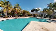 This single-story property is defined by its impressive outdoor amenity space, centered around a large, resort-style swimming pool with clear turquoise water and a spacious travertine or stone deck. The pool area is framed by an abundance of mature tropical palm trees and lush greenery, creating a private, verdant atmosphere reminiscent of a resort retreat. Lounge chairs and shaded umbrella seating are positioned throughout the deck, offering ample space for relaxation under the open blue sky. A well-maintained clubhouse or pavilion structure sits adjacent to the pool, featuring a low-pitched hip roof, large windows, and covered porch areas that provide a welcoming gathering space. The architecture of the building complements the tropical setting with its clean, classic design. The landscaping throughout the grounds is dense and carefully curated, with a variety of palms, shrubs, and potted plants adding layers of texture and color. The overall ambiance of the outdoor space is bright, open, and inviting, making it well-suited for leisure and social activities in a warm-weather environment. The interior spaces, while not fully illuminated in available imagery, appear to include additional functional rooms that support the overall use of the property.