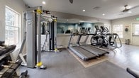 This fitness center is a fully outfitted, single-floor facility designed to accommodate a wide range of workout routines. The space is finished with a combination of wood-look luxury vinyl plank flooring in the open central area and protective rubber mat flooring beneath the weight and strength training equipment, providing both style and functionality.

The cardio zone features multiple treadmills and an elliptical trainer, lined along a large wall mirror that visually expands the space and allows users to monitor their form. A ceiling fan overhead supports ventilation and comfort during intense sessions. Adjacent to the cardio area, a dedicated strength training section houses a full power rack with an adjustable bench and weight plates, a multi-function cable machine, and a spin bike positioned in the open floor area.

The free weights section is well-stocked with a full rack of hex dumbbells spanning a wide weight range, a barbell weight tree, and a workout reference chart mounted on the wall for guidance. Natural light filters in through windows, brightening the weight room area. The walls are painted in a calm slate blue and grey palette, contributing to a clean and focused workout environment. Recessed lighting throughout ensures consistent illumination, and clearly marked doors provide convenient entry and exit access.