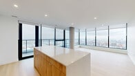 Located in Halifax, NS, this modern 2-bedroom, 3-bathroom residence offers a refined living experience defined by sleek design and breathtaking elevated views of the Halifax Harbour and cityscape. The open-concept kitchen and living area serves as the heart of the unit, anchored by a striking island with a warm wood-panelled base and crisp white quartz countertop. The kitchen features high-gloss white cabinetry, stainless steel appliances, and recessed lighting, all framed by expansive floor-to-ceiling windows that flood the space with natural light. The living area extends generously, with light hardwood flooring and panoramic corner windows offering unobstructed views in multiple directions. A clean, well-lit hallway with recessed lighting connects the living spaces to the private quarters. The spacious primary bedroom continues the floor-to-ceiling window theme, delivering stunning elevated views of the harbour and surrounding neighbourhood. Its ensuite features a wood-toned vanity with quartz counters and a large window overlooking the city. The additional bathrooms are finished in neutral stone-toned tile with glass-enclosed showers. Throughout, the design maintains a consistent modern aesthetic with white walls, light wood floors, and an abundance of natural light.