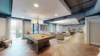 Located at 55 Romney St in Charleston's historic 29403 zip code, this thoughtfully designed amenity space offers a generous, open single-floor layout ideal for socializing, relaxing, and focused work. The interior is anchored by a striking herringbone hardwood floor that flows seamlessly throughout, lending warmth and visual continuity to the entire space.

The main gathering area features a full-size billiards table beneath a statement pendant light fixture, surrounded by ample room for movement. Large floor-to-ceiling windows with sheer drapery invite abundant natural light and offer views to the outdoors. A dedicated lounge zone provides a refined sitting area centered around a floor-to-ceiling stone-clad fireplace, complemented by upholstered armchairs and a plush area rug — perfect for relaxed conversation.

Exposed navy-painted ductwork adds an industrial-chic contrast against the otherwise clean white ceilings and walls, while dark steel beam accents reinforce the modern aesthetic. A kitchenette area equipped with white cabinetry, a microwave, and under-counter appliances supports casual entertaining. A semi-private area with a dark wood-paneled partition, marble-tile flooring, and built-in desk provides a quiet retreat for individual work. A bar-height dining counter rounds out the space's versatility.