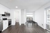 This contemporary studio unit is thoughtfully designed to maximize functionality within a compact, open-concept layout. The main living area features warm wood-tone laminate flooring and clean white walls, creating a bright, neutral backdrop. Large windows along one wall allow abundant natural light to fill the space while framing views of the surrounding urban neighborhood. A private balcony or exterior door provides additional outdoor access, enhancing the sense of openness.

The kitchen is positioned along one wall and is well-equipped with full stainless steel appliances, including a refrigerator, electric range, built-in microwave, and dishwasher. Crisp white cabinetry extends to the ceiling, offering ample storage, while light countertops and a tile backsplash complete the modern aesthetic. Recessed lighting illuminates the kitchen area efficiently.

The bathroom is finished in a clean, contemporary style with white cabinetry, an undermount sink with a white quartz-style countertop, chrome hardware, and wood-tone flooring consistent with the rest of the unit. A closet with a hanging rod is accessible from the bathroom area, providing convenient storage.

Overall, the unit presents a clean, modern aesthetic with consistent finishes throughout, making it a practical and well-appointed living space.